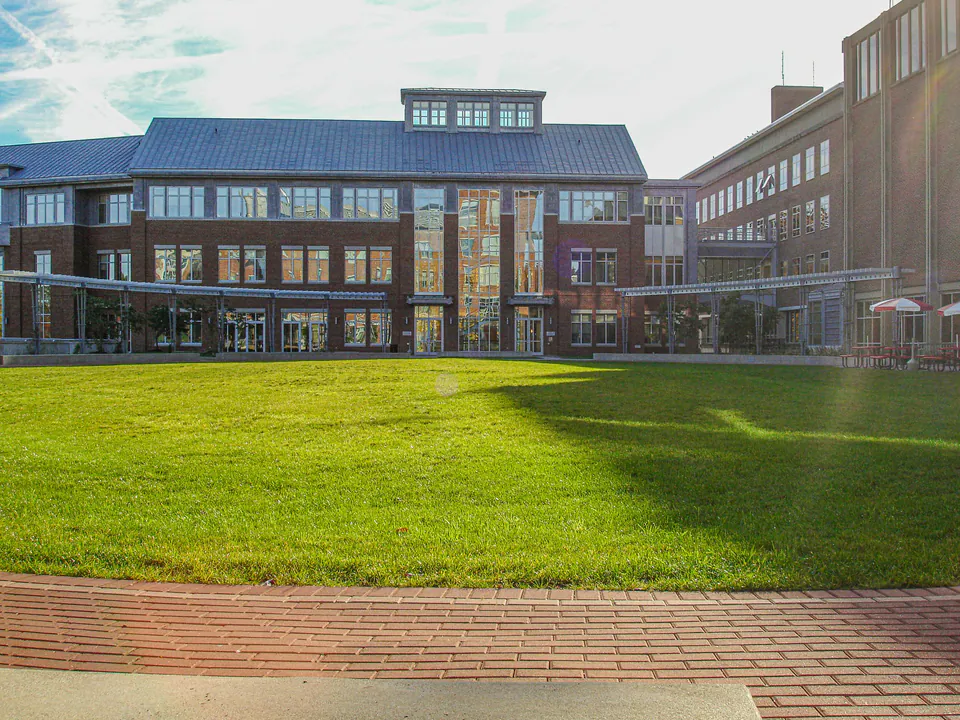 Campus building edge with low planting beds, young trees, brick walks, and concrete pedestrian surfaces